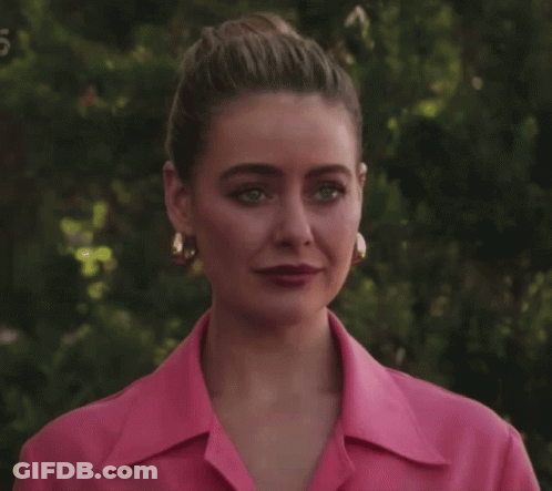 A Woman Wearing A Pink Shirt And Gold Hoop Earrings Stands In Front Of Trees GIF
