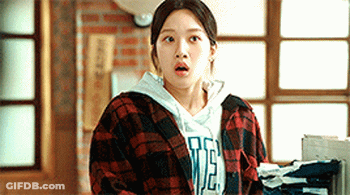 A Woman Wearing A Plaid Shirt And A Hoodie Is Standing In Front Of A Window . GIF