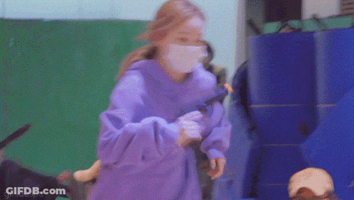 A Woman Wearing A Purple Hoodie And A White Mask Is Holding A Toy . GIF