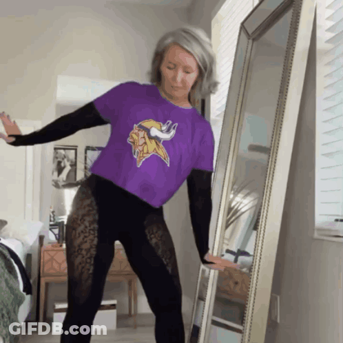 A Woman Wearing A Purple Minnesota Vikings Shirt Is Dancing In Front Of A Mirror GIF