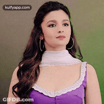A Woman Wearing A Purple Top And Hoop Earrings Is Standing In Front Of A Green Wall . GIF