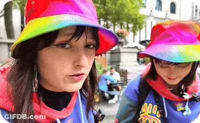 A Woman Wearing A Rainbow Hat Is Standing Next To Another Woman Wearing A Blue Shirt That Says ' Noodles ' GIF