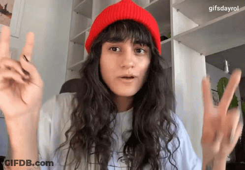 A Woman Wearing A Red Beanie And A White Shirt Is Making A Peace Sign GIF