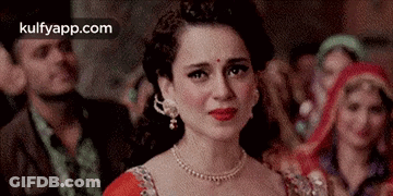 A Woman Wearing A Red Dress And A Necklace Is Crying In Front Of A Crowd . GIF
