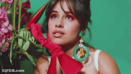 A Woman Wearing A Red Glove And Green Earrings Is Standing In Front Of A Vase Of Flowers GIF
