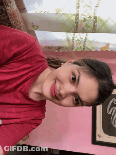 A Woman Wearing A Red Shirt Is Smiling In Front Of A Pink Wall GIF