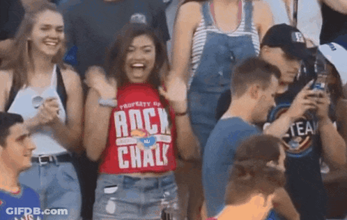A Woman Wearing A Red Shirt That Says Rock Chalk Stands In A Crowd Of People GIF