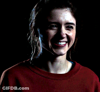 A Woman Wearing A Red Sweatshirt With A Pocket On The Front Smiles In The Dark GIF
