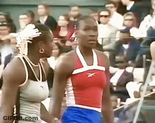A Woman Wearing A Red White And Blue Reebok Tank Top Stands Next To Another Woman GIF