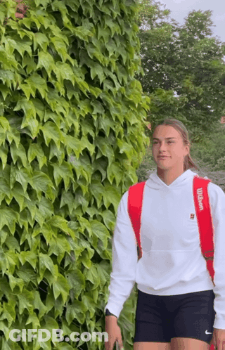 A Woman Wearing A Red Wilson Backpack Stands In Front Of A Lush Green Wall GIF