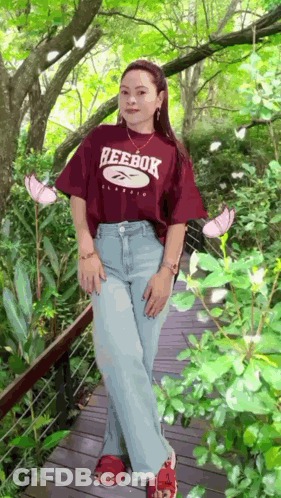 A Woman Wearing A Reebok T-Shirt Is Standing On A Wooden Bridge GIF