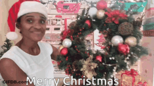 A Woman Wearing A Santa Hat Is Holding A Christmas Wreath In Front Of A Christmas Tree GIF
