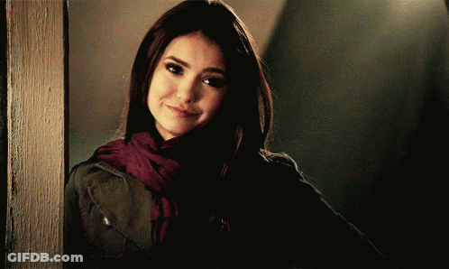 A Woman Wearing A Scarf And A Jacket Is Standing Next To A Wall GIF
