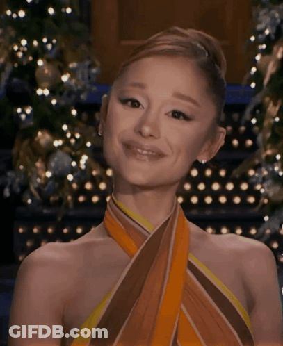 A Woman Wearing A Scarf Around Her Neck Is Smiling In Front Of A Christmas Tree . GIF