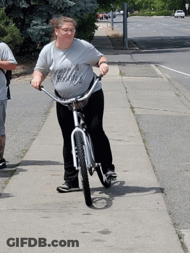 A Woman Wearing A Shirt That Says Cruise & Booze Is Riding A Bike Down A Sidewalk GIF