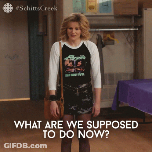 A Woman Wearing A Shirt That Says " What Are We Supposed To Do Now " Stands In A Room GIF