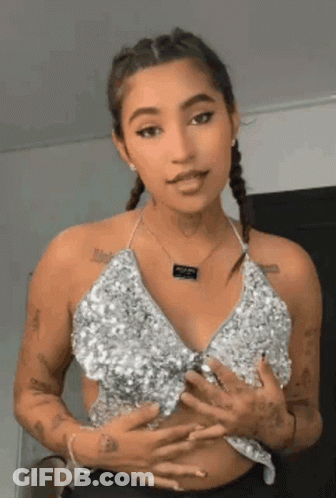 A Woman Wearing A Silver Top And A Necklace Is Standing In Front Of A Wall . GIF