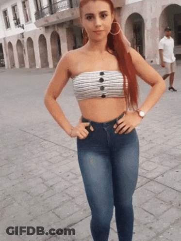 A Woman Wearing A Strapless Top And Jeans Stands On A Sidewalk With Her Hands On Her Hips GIF