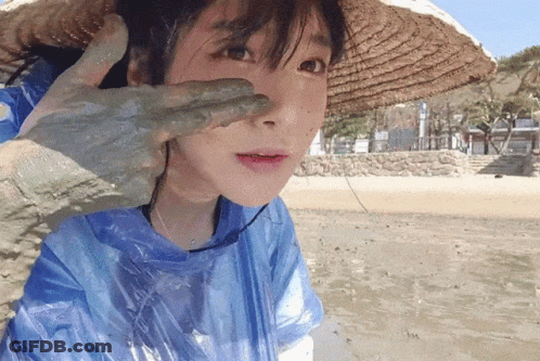 A Woman Wearing A Straw Hat And A Blue Raincoat Has A Hand Covered In Mud On Her Face GIF