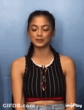 A Woman Wearing A Striped Crop Top And Jeans Is Sitting In Front Of A Blue Wall . GIF