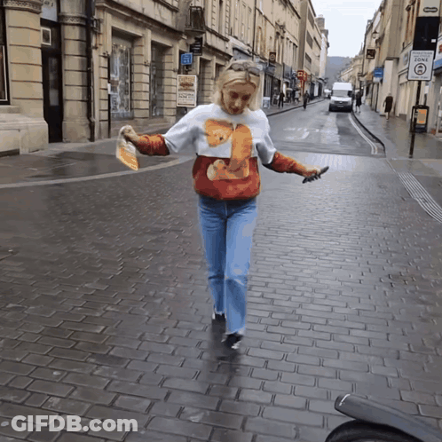 A Woman Wearing A Sweater With A Bear On It Is Walking Down A Brick Street GIF