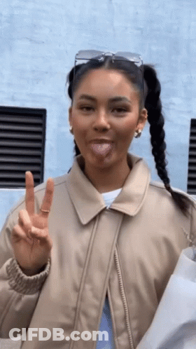 A Woman Wearing A Tan Jacket Is Sticking Her Tongue Out And Giving A Peace Sign GIF