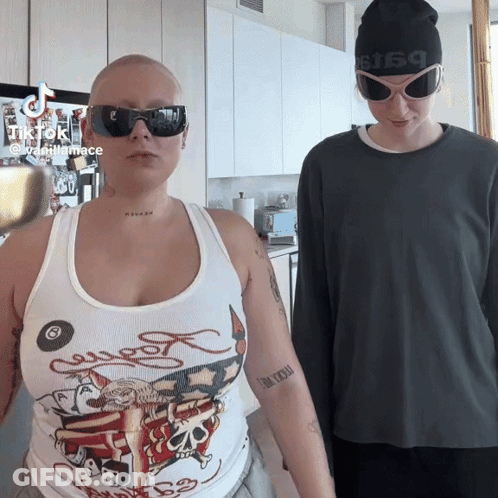 A Woman Wearing A Tank Top That Says Ed Hardy Is Standing Next To A Man Wearing Sunglasses GIF