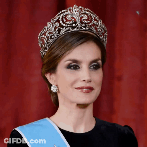 A Woman Wearing A Tiara And A Blue Sash Looks At The Camera GIF