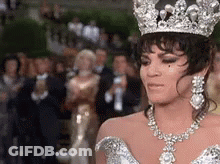 A Woman Wearing A Tiara And A Necklace Is Standing In Front Of A Crowd Of People . GIF