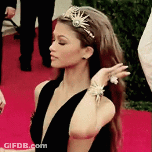 A Woman Wearing A Tiara And Bracelets Is Standing On A Red Carpet GIF