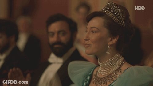 A Woman Wearing A Tiara And Pearls Sits In Front Of A Hbo Sign GIF
