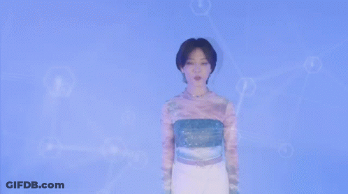 A Woman Wearing A Tie Dye Shirt And A Necklace Looks At The Camera . GIF