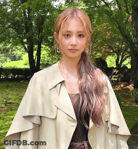 A Woman Wearing A Trench Coat And Earrings Is Standing In A Park GIF