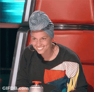 A Woman Wearing A Turban And Earrings Smiles While Sitting In A Chair GIF
