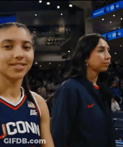 A Woman Wearing A Uconn Jersey Stands Next To A Woman Wearing A Nike Jacket GIF