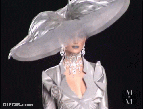 A Woman Wearing A Very Large Hat And A Necklace With The Letter M On The Bottom GIF