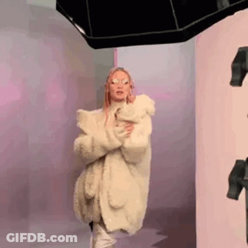 A Woman Wearing A White Fur Coat And Sunglasses Is Standing In Front Of A Mirror . GIF