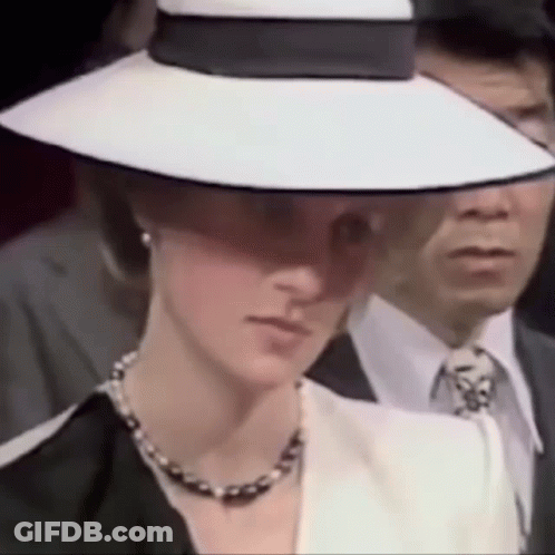 A Woman Wearing A White Hat And A Black Jacket Is Standing Next To A Man In A Suit . GIF