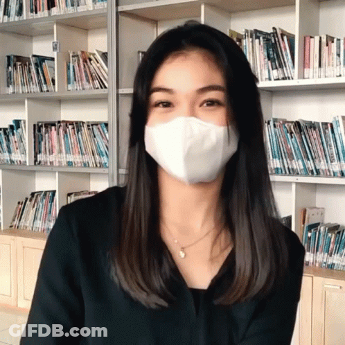A Woman Wearing A White Mask Stands In Front Of A Book Shelf GIF