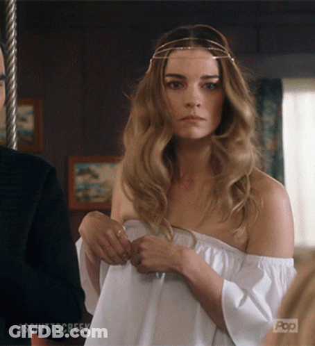 A Woman Wearing A White Off The Shoulder Top Is Standing Next To A Man With #Schittscreek On The Bottom Right GIF