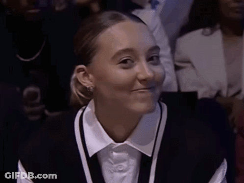 A Woman Wearing A White Shirt And A Black Vest Smiles While Sitting In A Crowd GIF