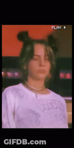 A Woman Wearing A White Shirt And A Necklace Looks At The Camera With A Play Button In The Corner GIF