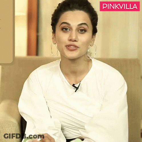 A Woman Wearing A White Shirt And Hoop Earrings Sits In Front Of A Pinkvilla Logo GIF