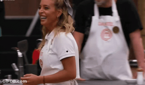 A Woman Wearing A White Shirt With A Heart On It Is Smiling In Front Of A Man Wearing An Apron GIF