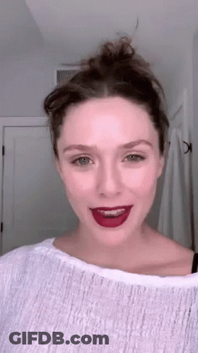 A Woman Wearing A White Sweater And Red Lipstick Is Standing In Front Of A Mirror . GIF
