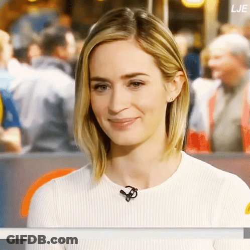 A Woman Wearing A White Sweater With A Microphone Around Her Neck Looks At The Camera GIF