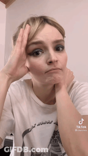 A Woman Wearing A White T-Shirt Has Her Hand On Her Forehead . GIF
