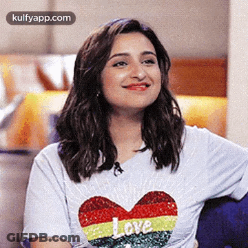 A Woman Wearing A White T-Shirt With A Heart On It Is Smiling And Sitting On A Couch . GIF