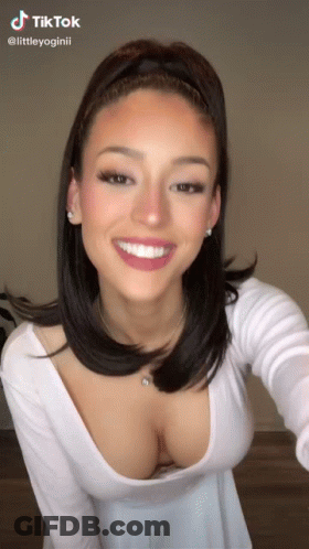 A Woman Wearing A White Top With A Plunging Neckline Is Smiling . GIF