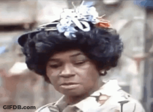 A Woman Wearing A Wig And A Hat With Flowers On It Is Making A Funny Face . GIF